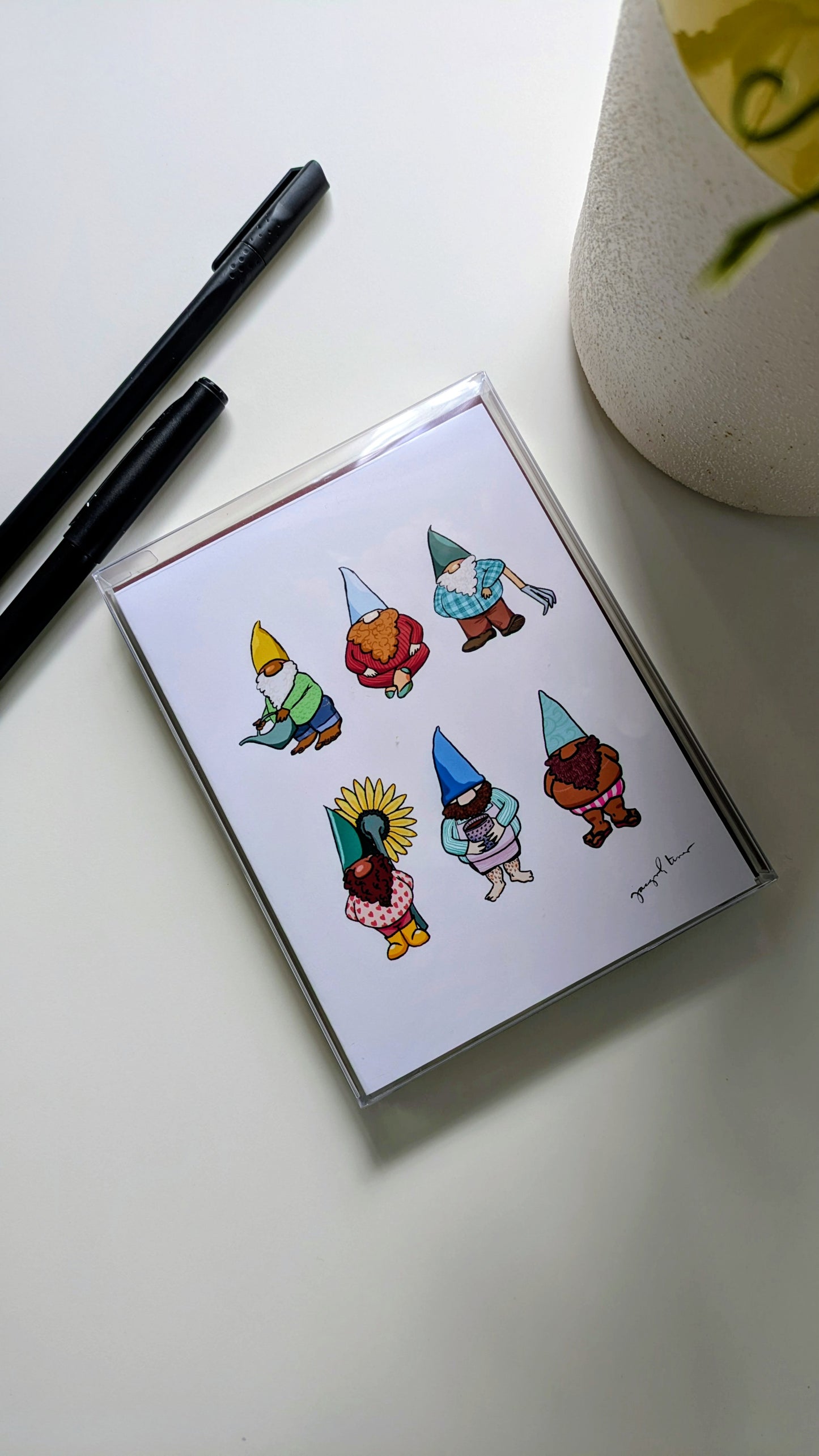 A light purple notecard set with burgundy envelopes is nestled inside a clear plastic box on a white background, surrounded by pens. The notecard shows six gnomes arranged in a grid. Some gnomes are holding gardening tools, others are holding flowers. Each gnome has a different skin tone.