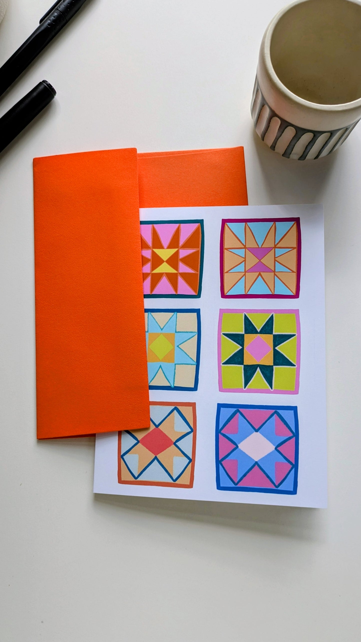 A single notecard nestled in a bright orange envelope, surrounded by pens on a white background. The notecard shows six brightly colored square quilt blocks arranged in a grid.
