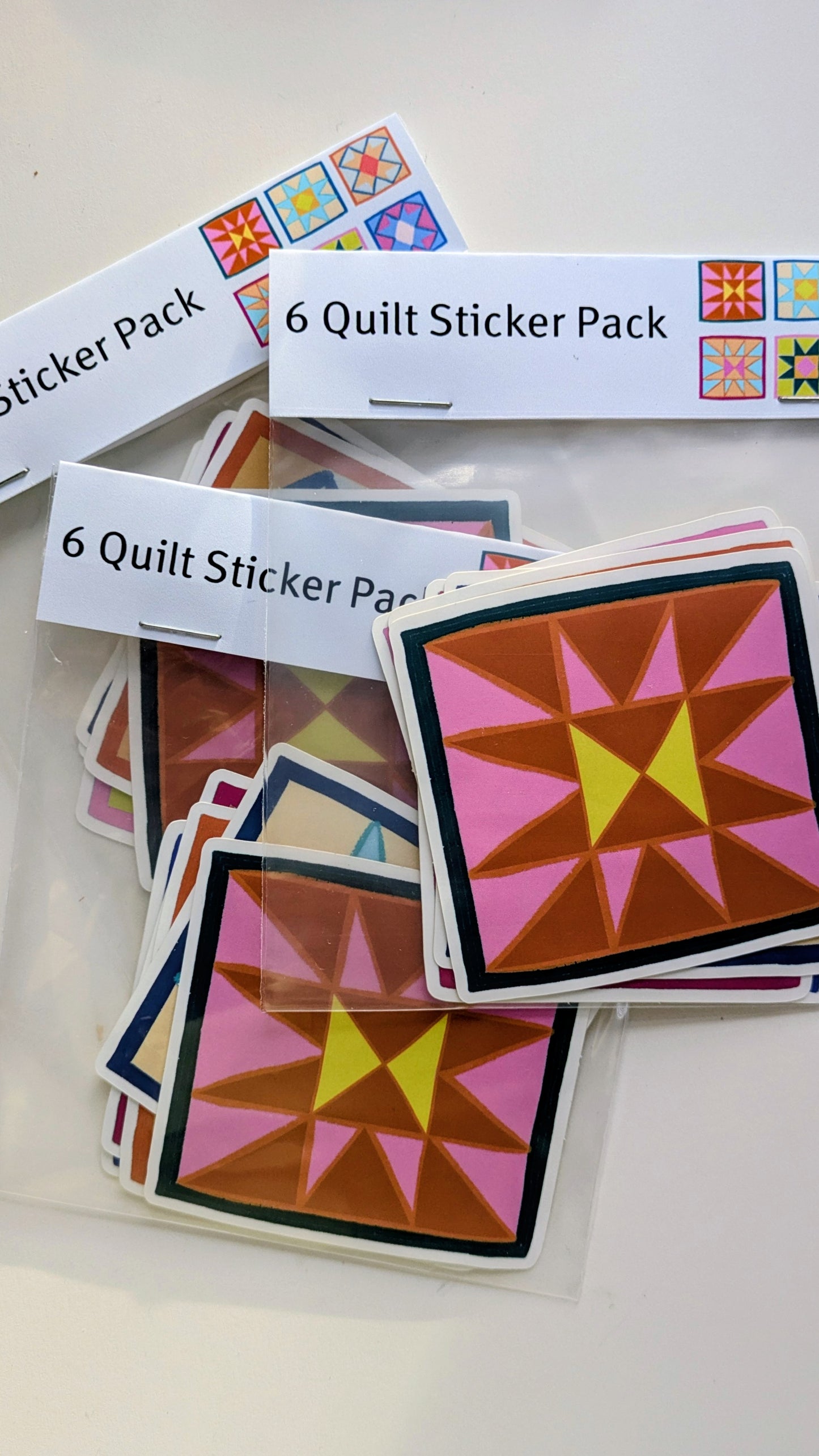 Multiple sticker packs lying on a white background, each showing a half-square triangle quilt block in pink, orange and yellow.