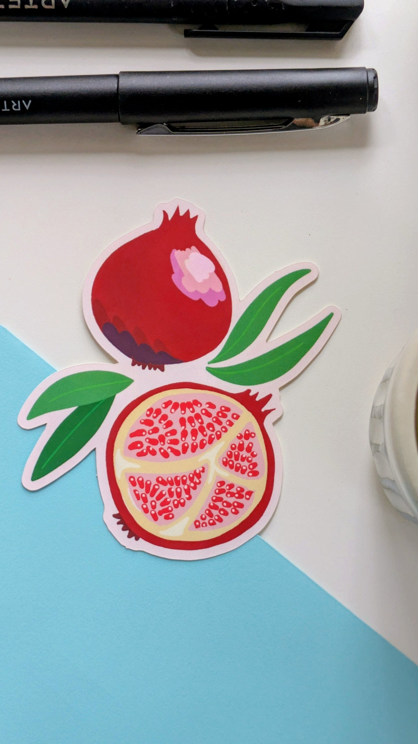 Image of a sticker of two pomegranates, one whole and one split in half, with leaves reaching out between them. The sticker rests atop a white background and a bright blue envelope.