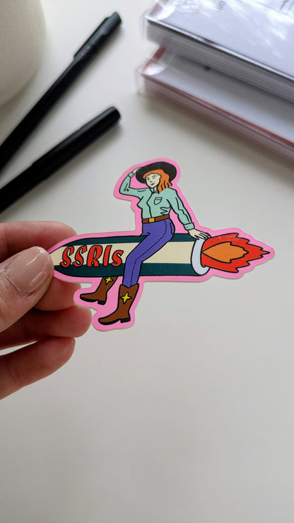 Image of a vinyl sticker being held by a hand over a white background. The sticker features light-skinned cowgirl with red hair riding a fiery green and white rocket with “SSRIs” written on it.