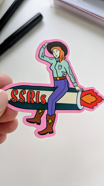 Image of a vinyl sticker being held by a hand over a white background. The sticker features light-skinned cowgirl with red hair riding a fiery green and white rocket with “SSRIs” written on it.