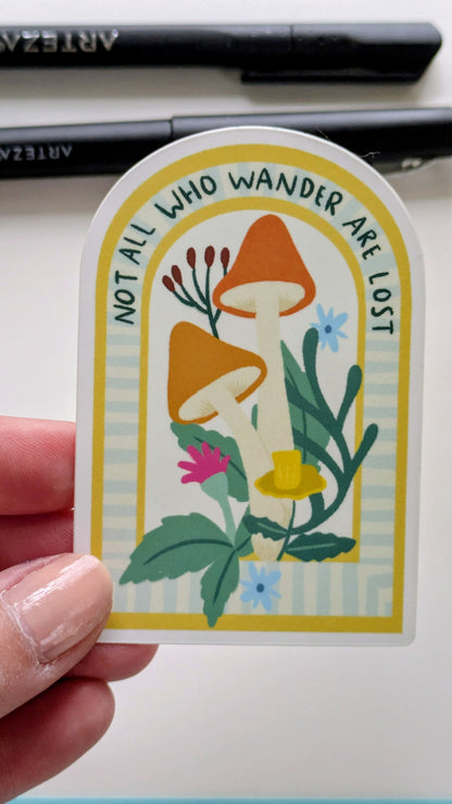 Light-skinned hand holds a sticker of mushrooms and flowers nestled in an archway, with the words “Not all who wander are lost” written along the top of the arch.