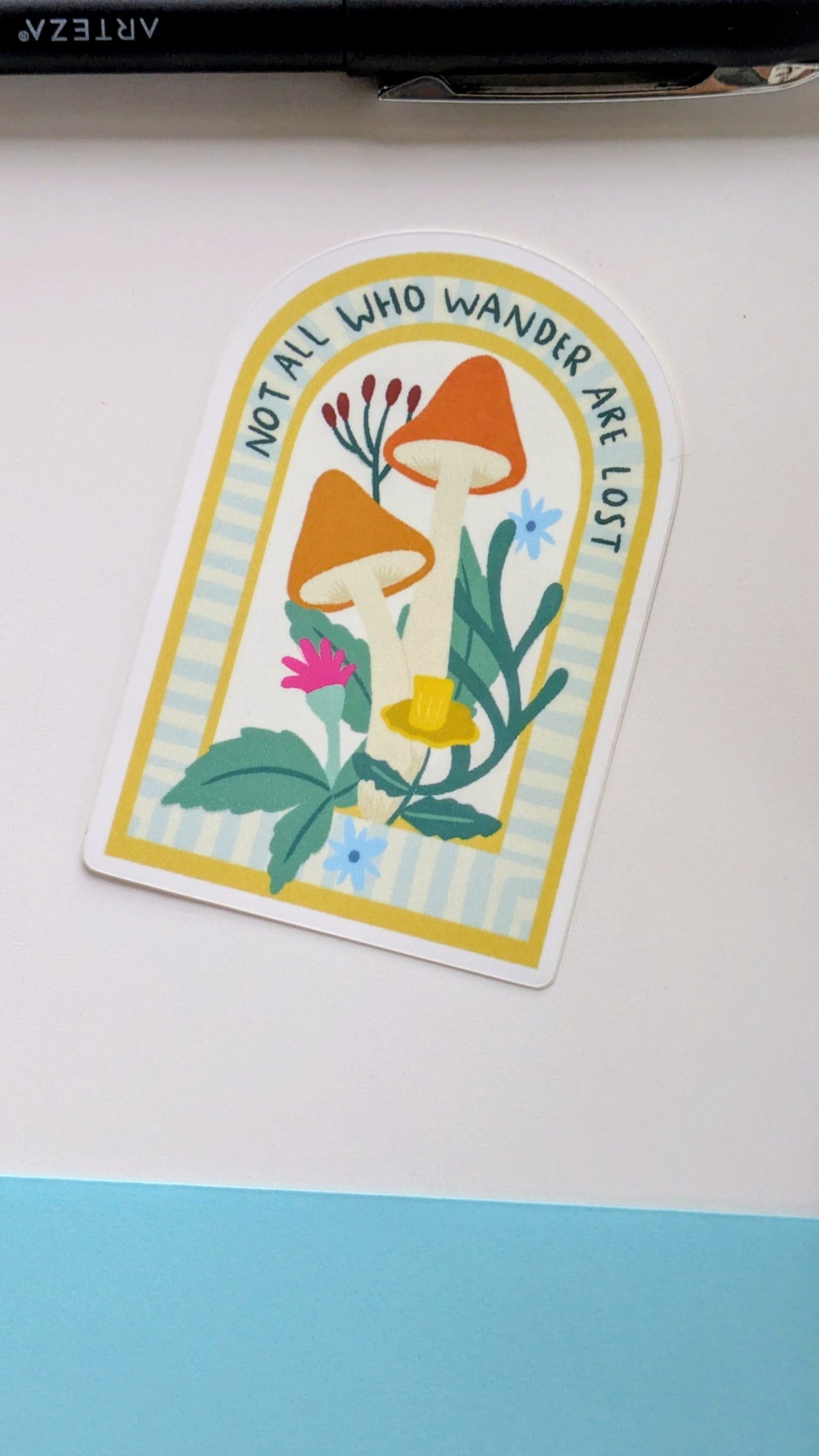 On a white background lies a sticker of mushrooms and flowers nestled in an archway, with the words “Not all who wander are lost” written along the top of the arch.