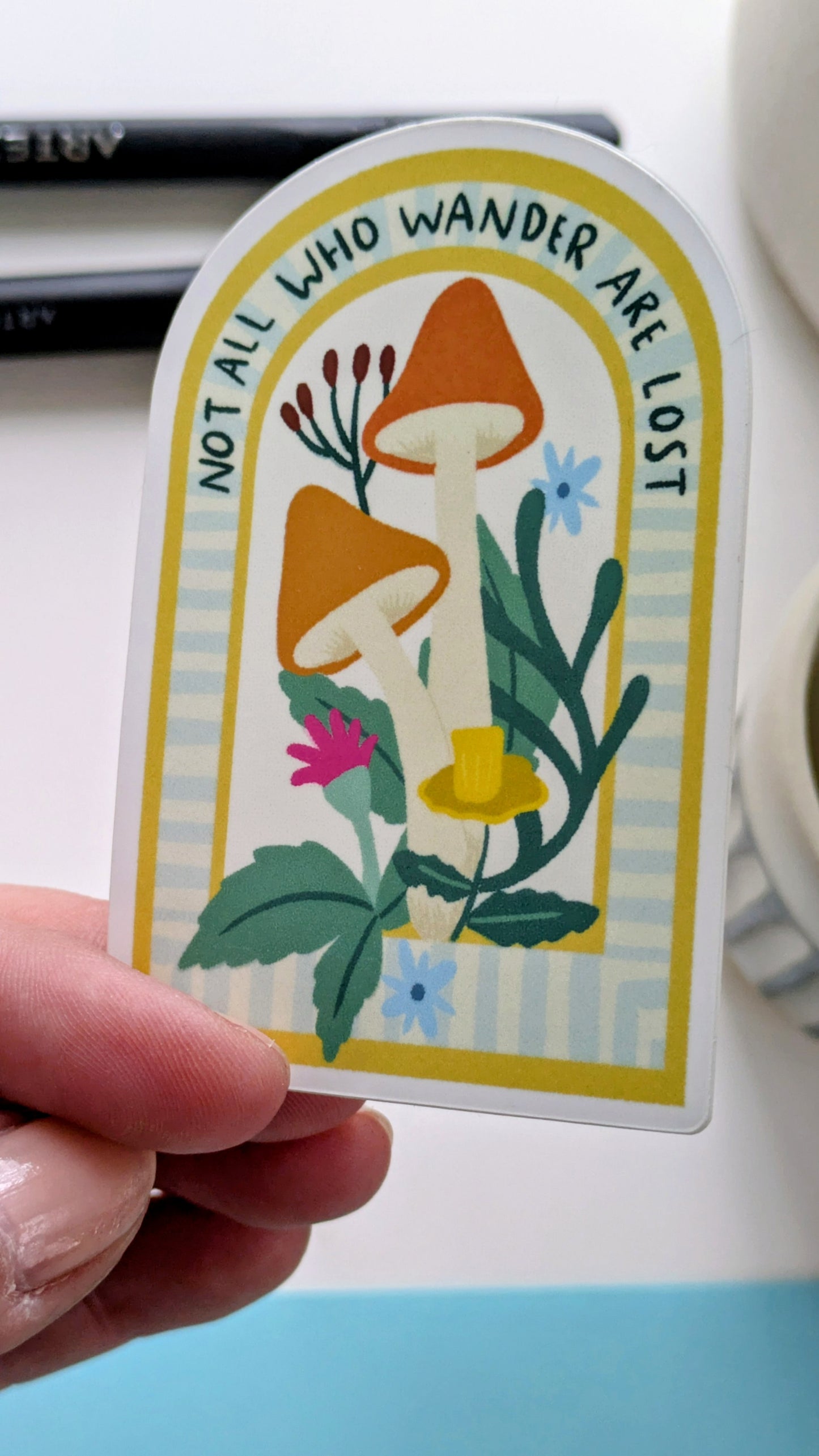 A Light-skinned hand holds a sticker of mushrooms and flowers nestled in an archway, with the words “Not all who wander are lost” written along the top of the arch.