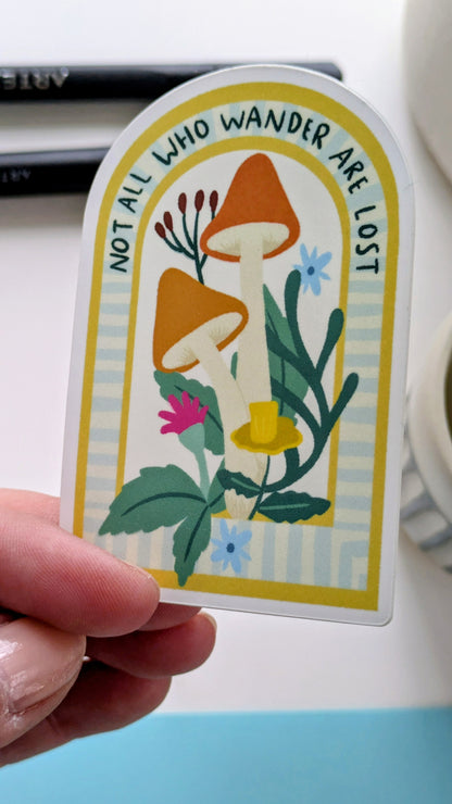A Light-skinned hand holds a sticker of mushrooms and flowers nestled in an archway, with the words “Not all who wander are lost” written along the top of the arch.