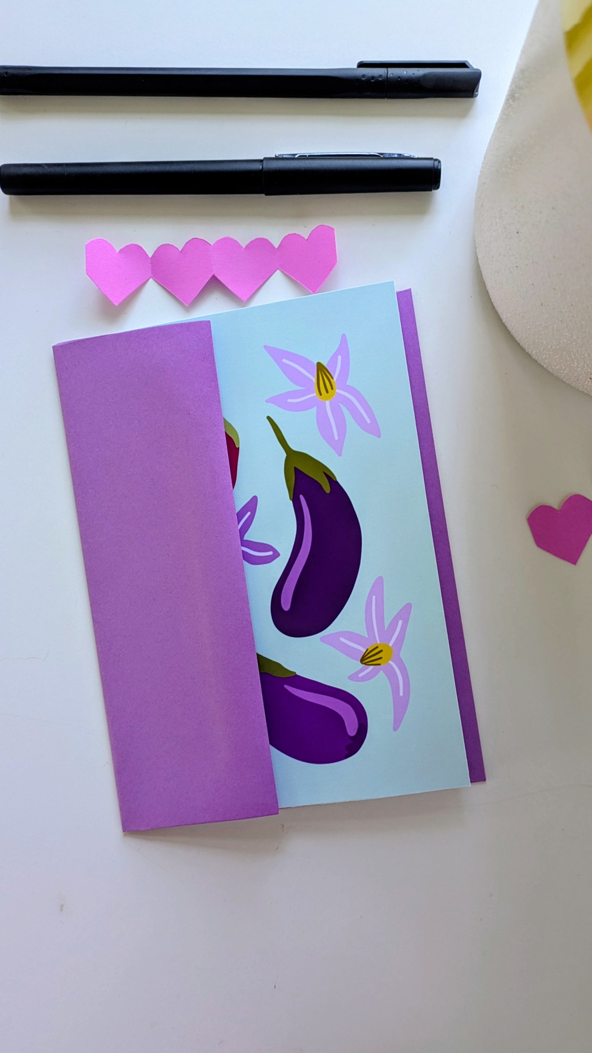 Soft green card sits inside of a purple envelope, surrounded by paper cut-out hearts and pens on top of a white background. The card features four eggplants of different sizes and colors, accompanied by purple flowers.