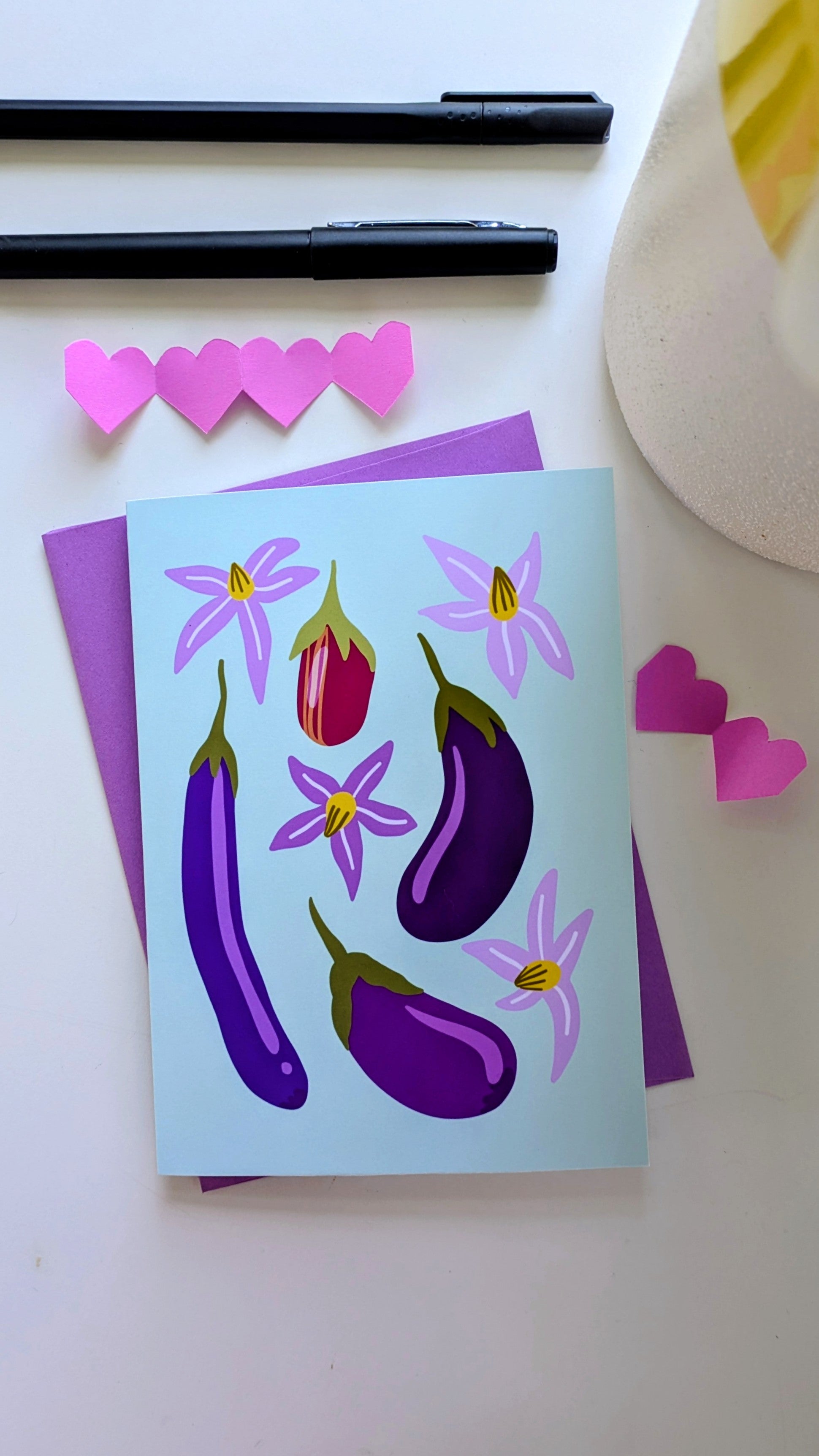 Soft green card sits on top of a purple envelope, surrounded by paper cut-out hearts and pens on top of a white background. The card features four eggplants of different sizes and colors, accompanied by purple flowers.