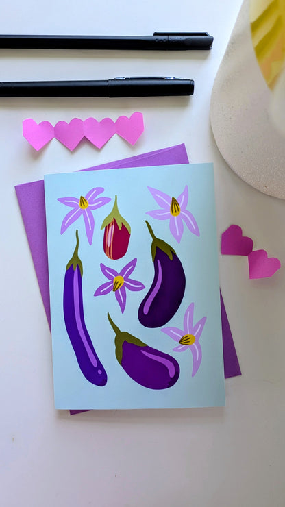 Soft green card sits on top of a purple envelope, surrounded by paper cut-out hearts and pens on top of a white background. The card features four eggplants of different sizes and colors, accompanied by purple flowers.