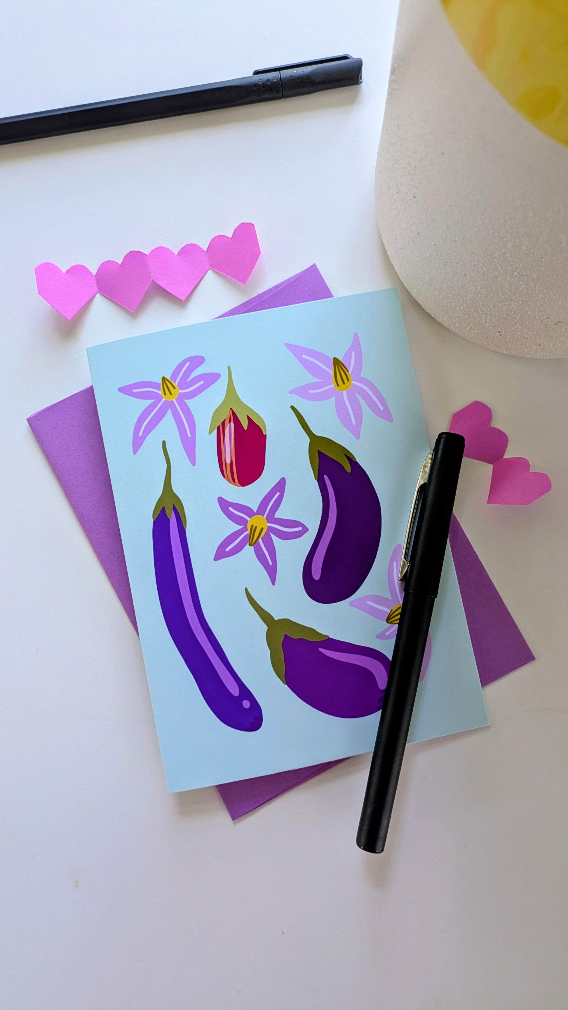 Soft green card sits on top of a purple envelope, surrounded by paper cut-out hearts and pens on top of a white background. The card features four eggplants of different sizes and colors, accompanied by purple flowers.