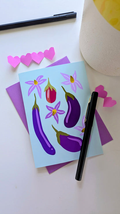 Soft green card sits on top of a purple envelope, surrounded by paper cut-out hearts and pens on top of a white background. The card features four eggplants of different sizes and colors, accompanied by purple flowers.