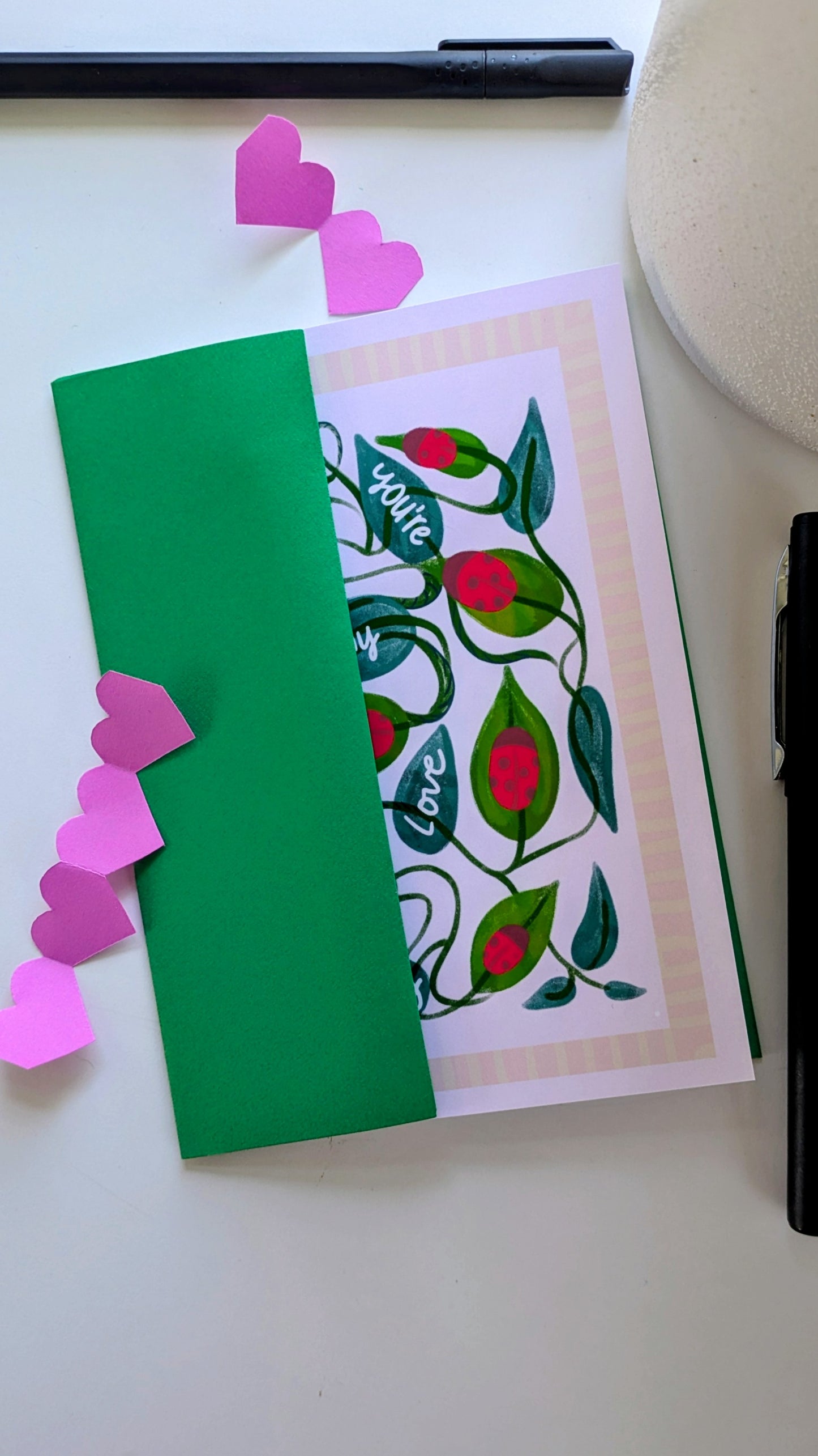 Card laying inside a green envelope, surrounded by paper cut-out hearts and pens. The card features twisting vines, leaves, and ladybugs. Four of the leaves have the words “you’re my love bug” written on them in white script. The image is surrounded by a pink geometrical border.