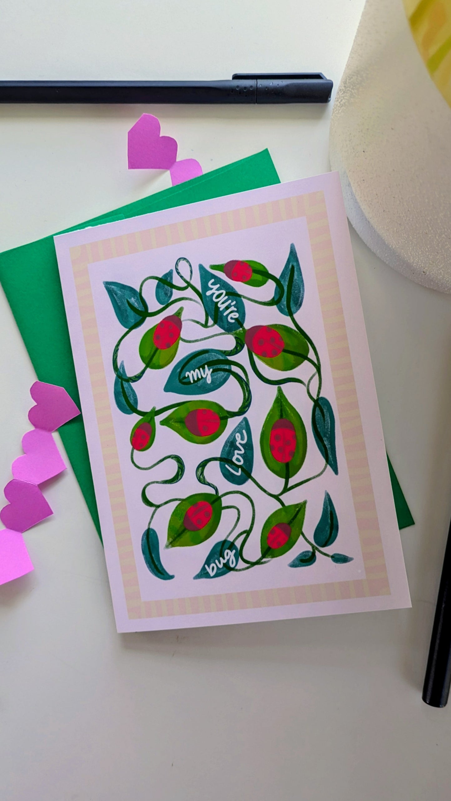 Card laying on a green envelope, surrounded by paper cut-out hearts and pens. The card features twisting vines, leaves, and ladybugs. Four of the leaves have the words “you’re my love bug” written on them in white script. The image is surrounded by a pink geometrical border.