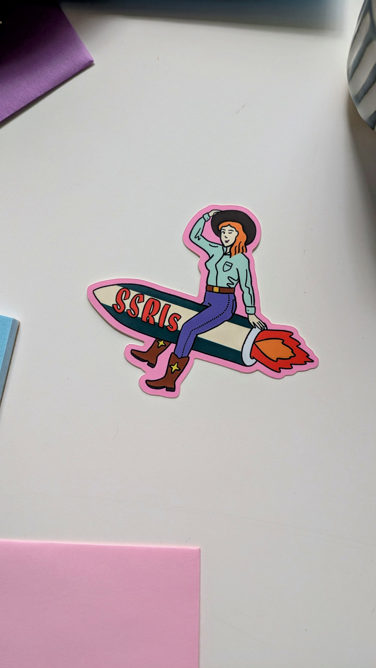 Image of a vinyl sticker on a white background. The sticker features light-skinned cowgirl with red hair riding a fiery green and white rocket with “SSRIs” written on it.