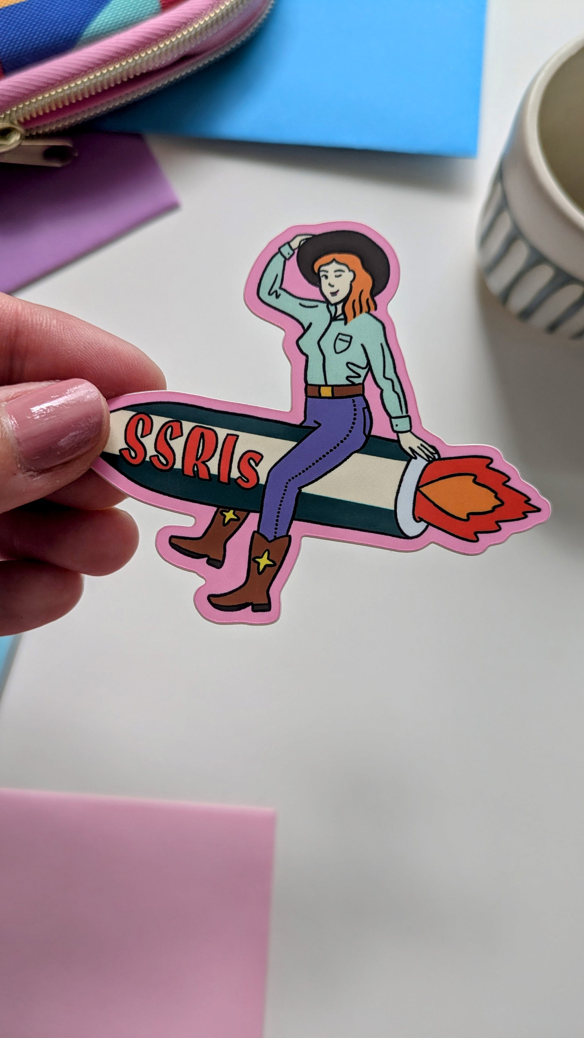 Image of a vinyl sticker being held by a hand over a white background. The sticker features light-skinned cowgirl with red hair riding a fiery green and white rocket with “SSRIs” written on it.